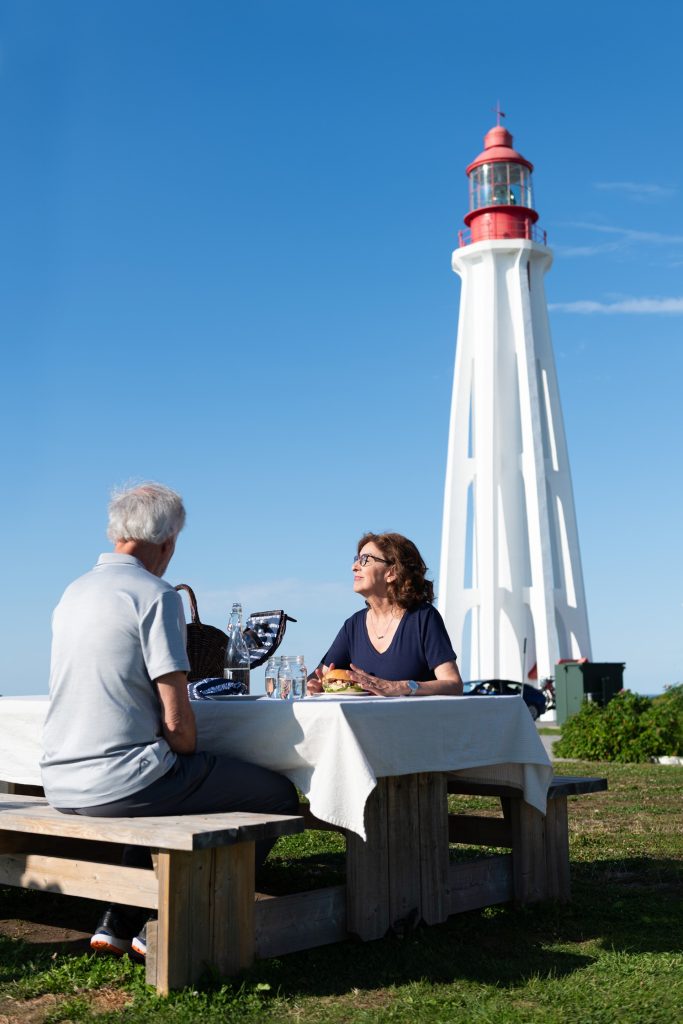 Phare de Pointe-au-Père avec des personnes à une table Site historique maritime de la Pointe-au-Père Crédit SHMP Phare de Pointe-au-Père avec des personnes à une table Site historique maritime de la Pointe-au-Père Crédit SHMP